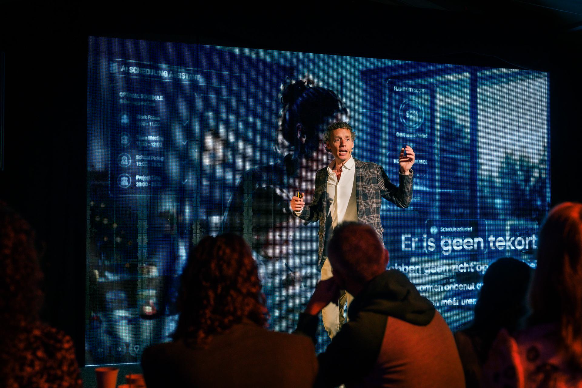 Laurens Waling speaking energetically to the room, with the words 'Er is geen tekort' on screen behind him