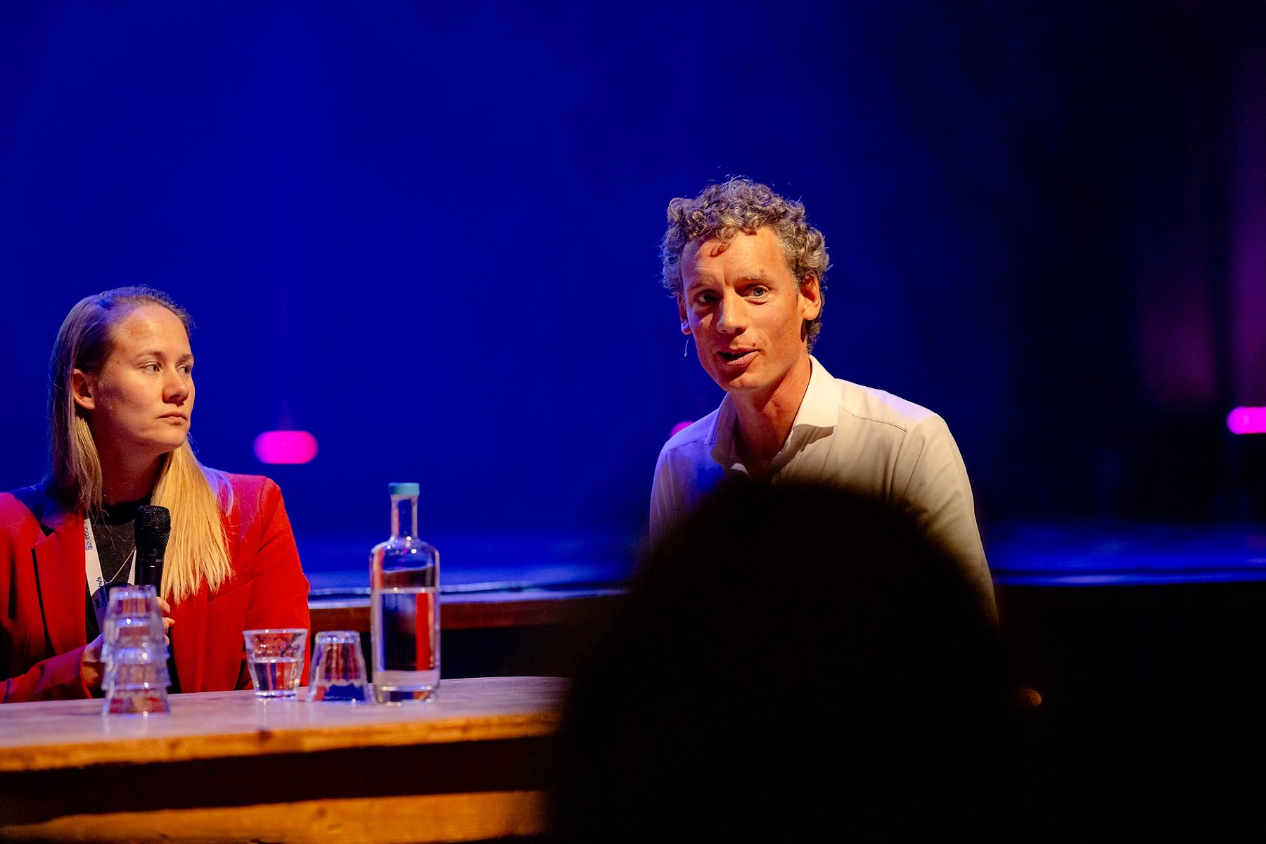 Laurens Waling in gesprek aan tafel op een blauw verlicht podium, naast een panellid dat luistert